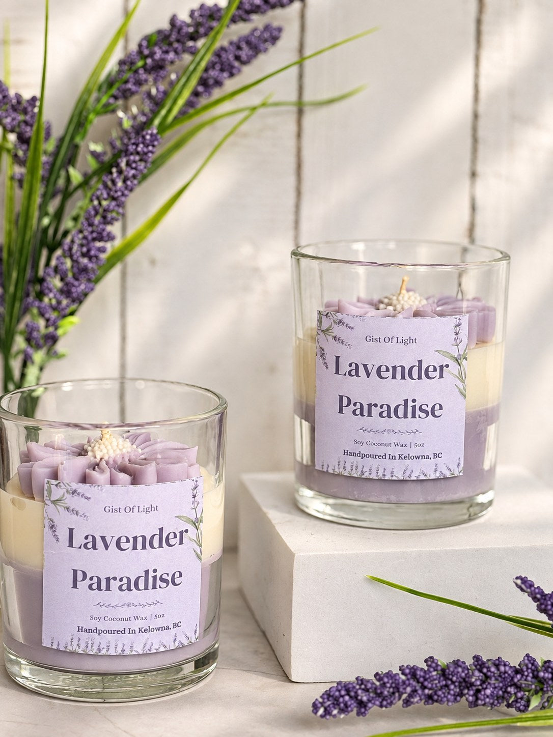 Two Lavender Paradise botanical candles on a white plinth with fresh lavender stems