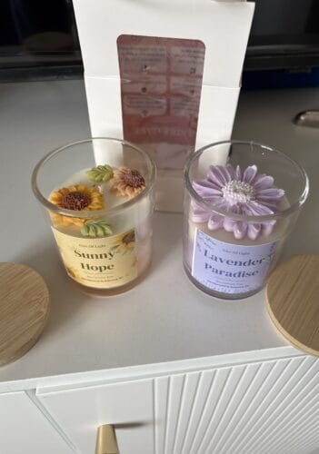 Two botanical floral candles in glass jars with flower decorations, on a white surface.