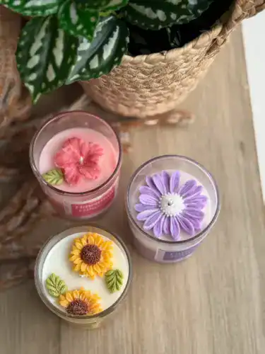 Colorful floral candles with green leaves in glass containers on wooden surface.