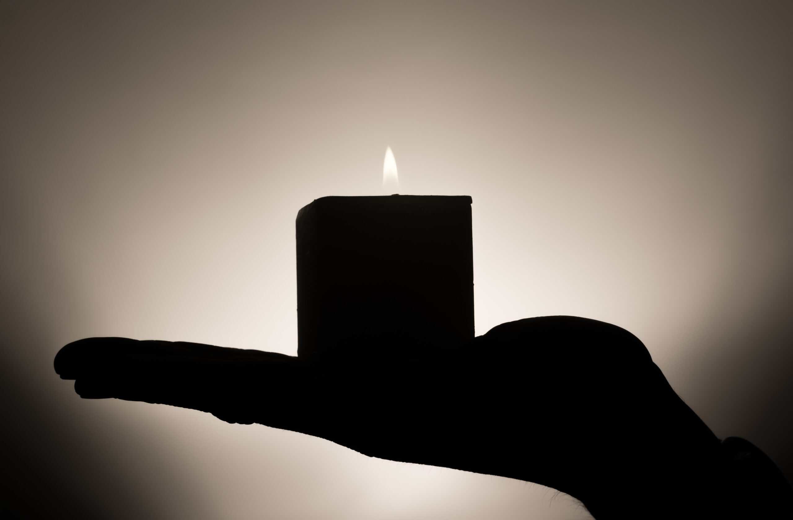 Silhouette of a hand holding a lit candle against a soft background.